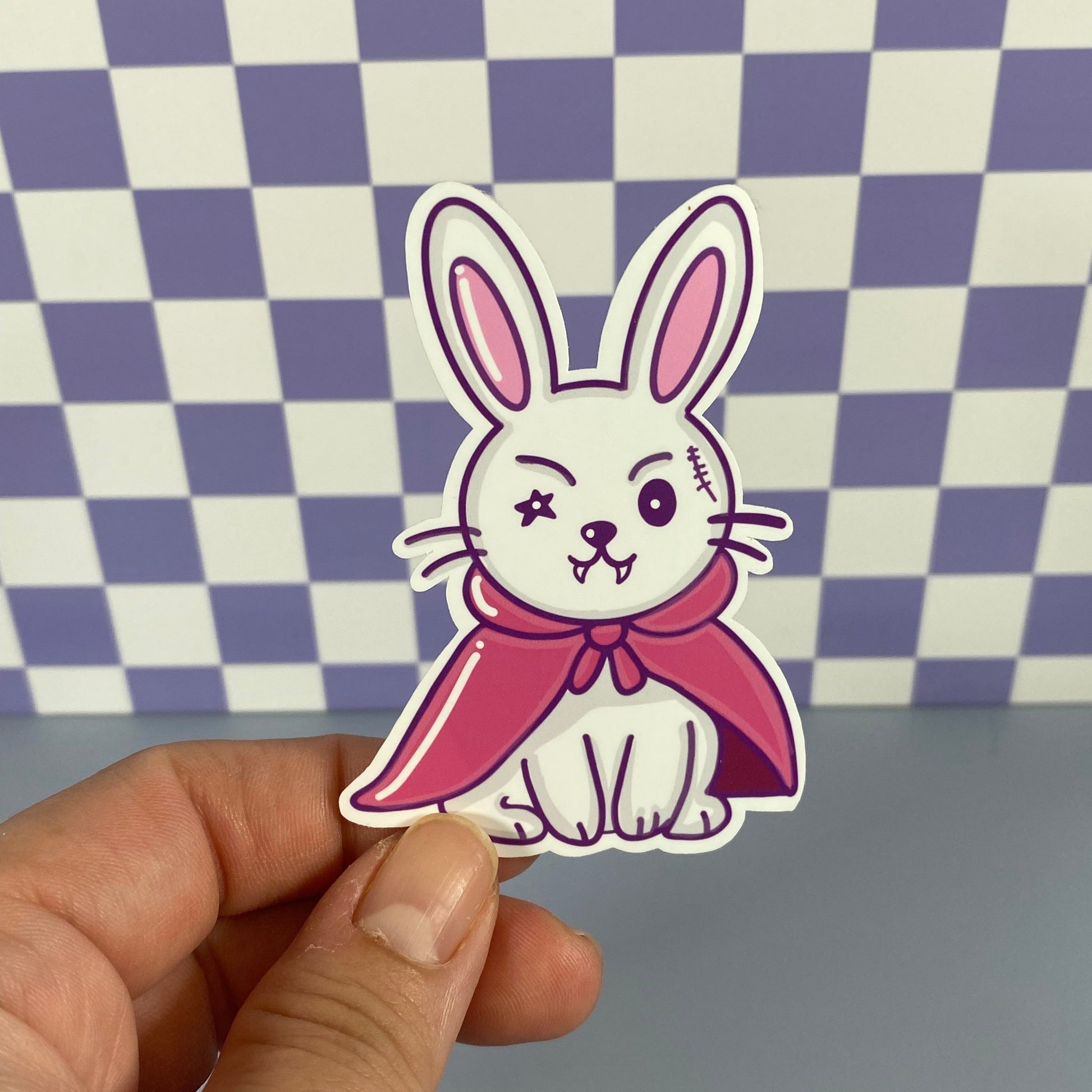 Hand holding a sticker of a rabbit with a pink cape against a checkered background